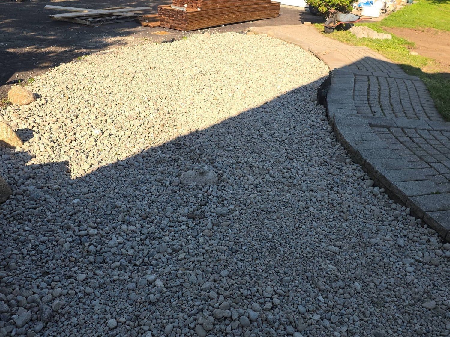 Fresh gravel beds meeting a curved interlock walkway with lumber staged for construction
